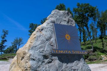 Large rock with a metal sign showing a sunburst logo and the words TENUTA STELLA against a blue sky at an outdoor site.