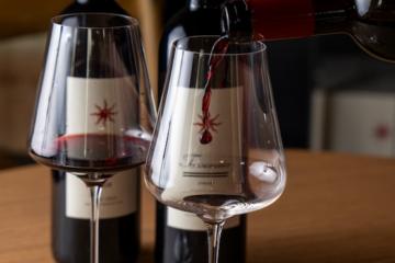 Two wine glasses on a wooden table, with red wine being poured into one glass from a bottle behind them.